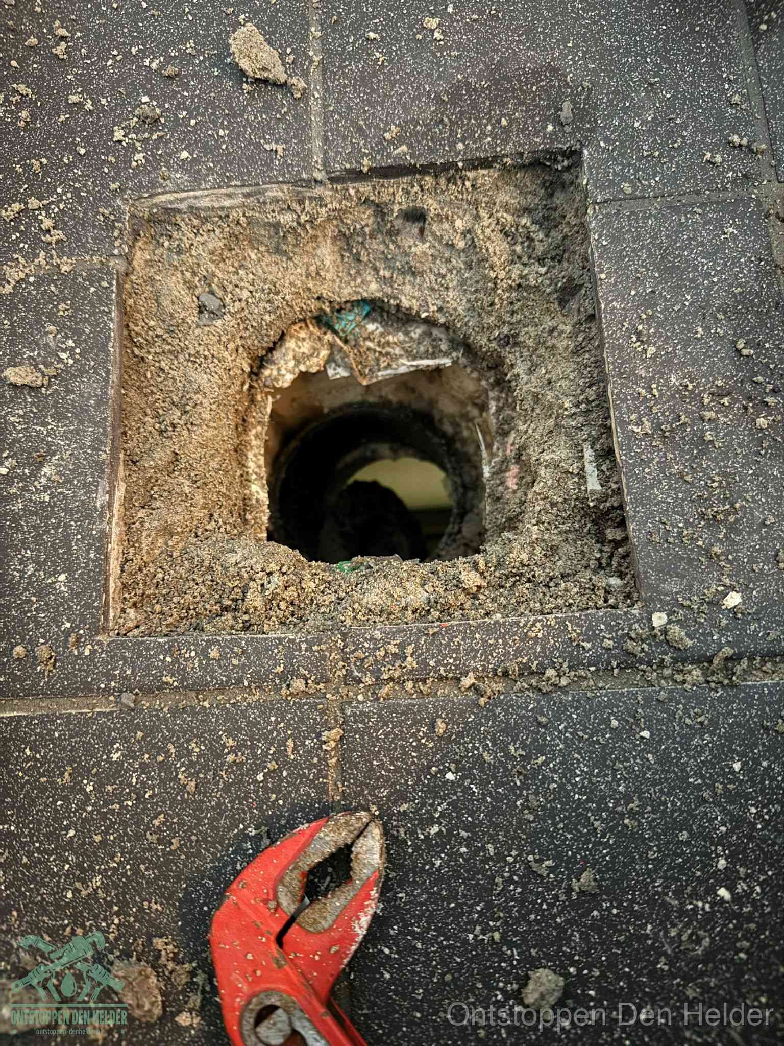 Een geopend rioolputje in een betontegelvloer met losgemaakt rooster en een zichtbare ronde afvoerbuis, het achterhalen waar de afvoerverstopping zit in Den Helder