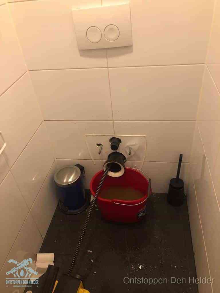 Een ontstopte wand-wc met verwijderde toiletpot waar een ontstoppingsveer in de wanduitgang verdwijnt, het achterhalen waar de verstopping zit in Den Helder