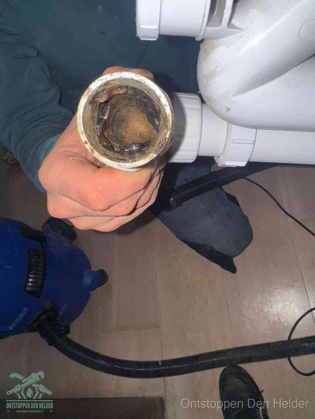Een hand houdt een losgekoppeld PVC-afvoerbuisstuk vast dat van binnen volledig is dichtgeslibd met vet, vuil en aanslag, de meest voorkomende oorzaak van een verstopte afvoer in Den Helder