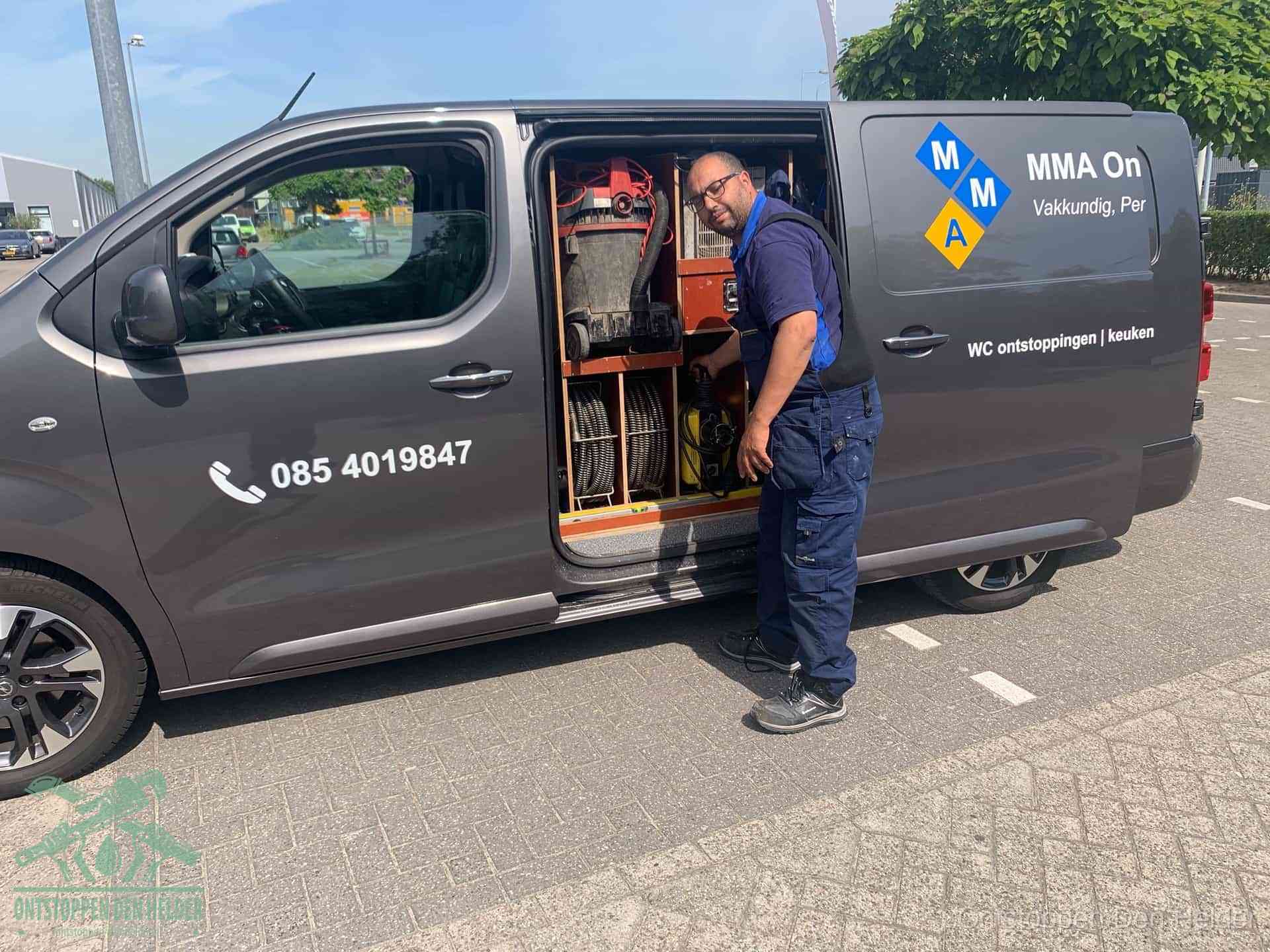 Een monteur bij de open schuifdeur van zijn bedrijfsbus op een parkeerplaats, klaar om de oorzaak van terugkerende afvoerverstoppingen aan te pakken in Den Helder