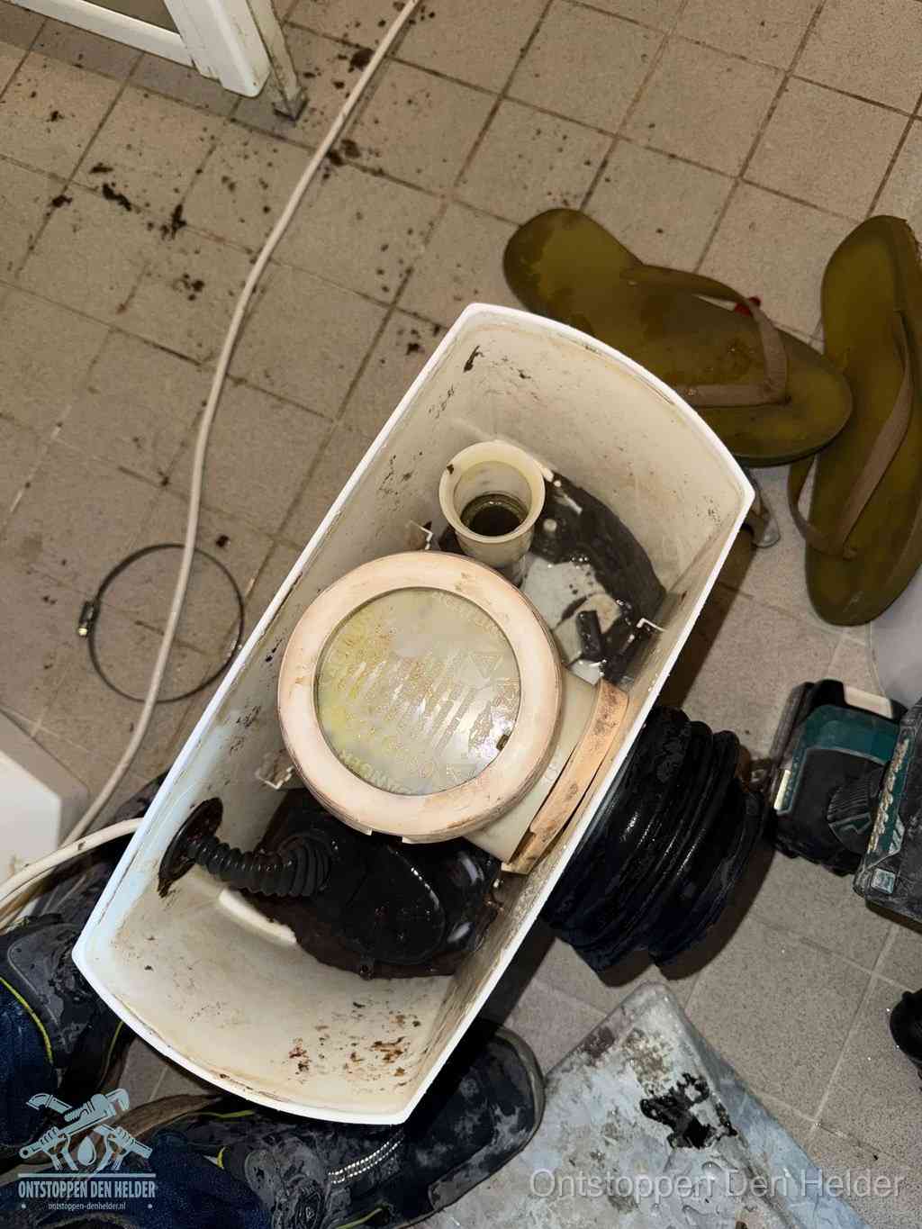 Een gedemonteerde toiletpot op de vloer met ontstoppingsveer en gereedschap ernaast, het verwijderen van een vastzittend voorwerp uit de wc in Den Helder