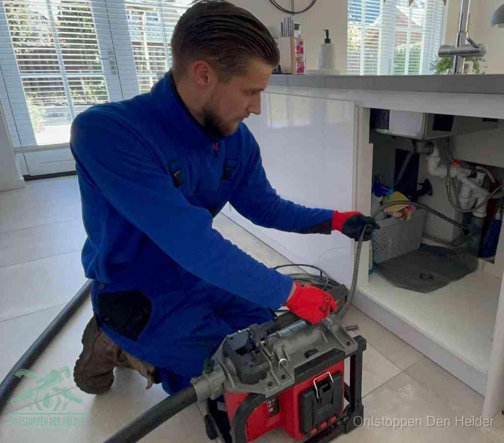Een monteur met rode handschoenen knielt in een keuken en voert een elektrische ontstoppingsveer in tijdens het professioneel afvoer ontstoppen in Den Helder