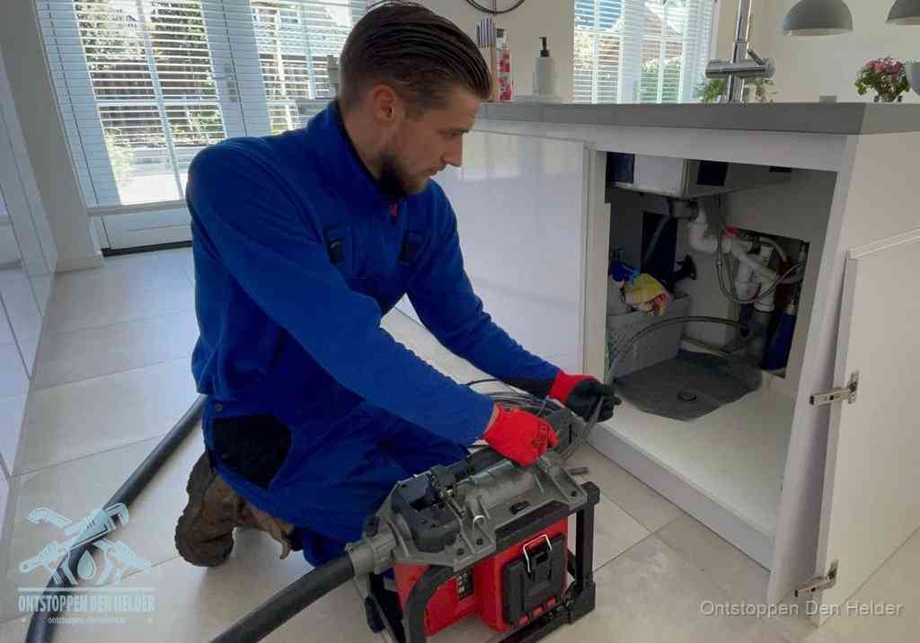 Een monteur voert een professionele elektrische ontstoppingsmachine in de afvoer onder de spoelbak tijdens het gootsteen ontstoppen in Den Helder