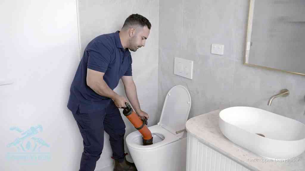 Een man gebruikt een handmatige luchtdruk-ontstopper in een modern betegeld toilet om zelf de wc te ontstoppen in Den Helder